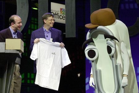Bill Gates presents a T-shirt as a retirement gift to "Clippy," a person dressed as the Microsoft Office Assistant, as Jeff Bezos looks on