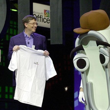 Bill Gates presents a T-shirt as a retirement gift to "Clippy," a person dressed as the Microsoft Office Assistant, as Jeff Bezos looks on