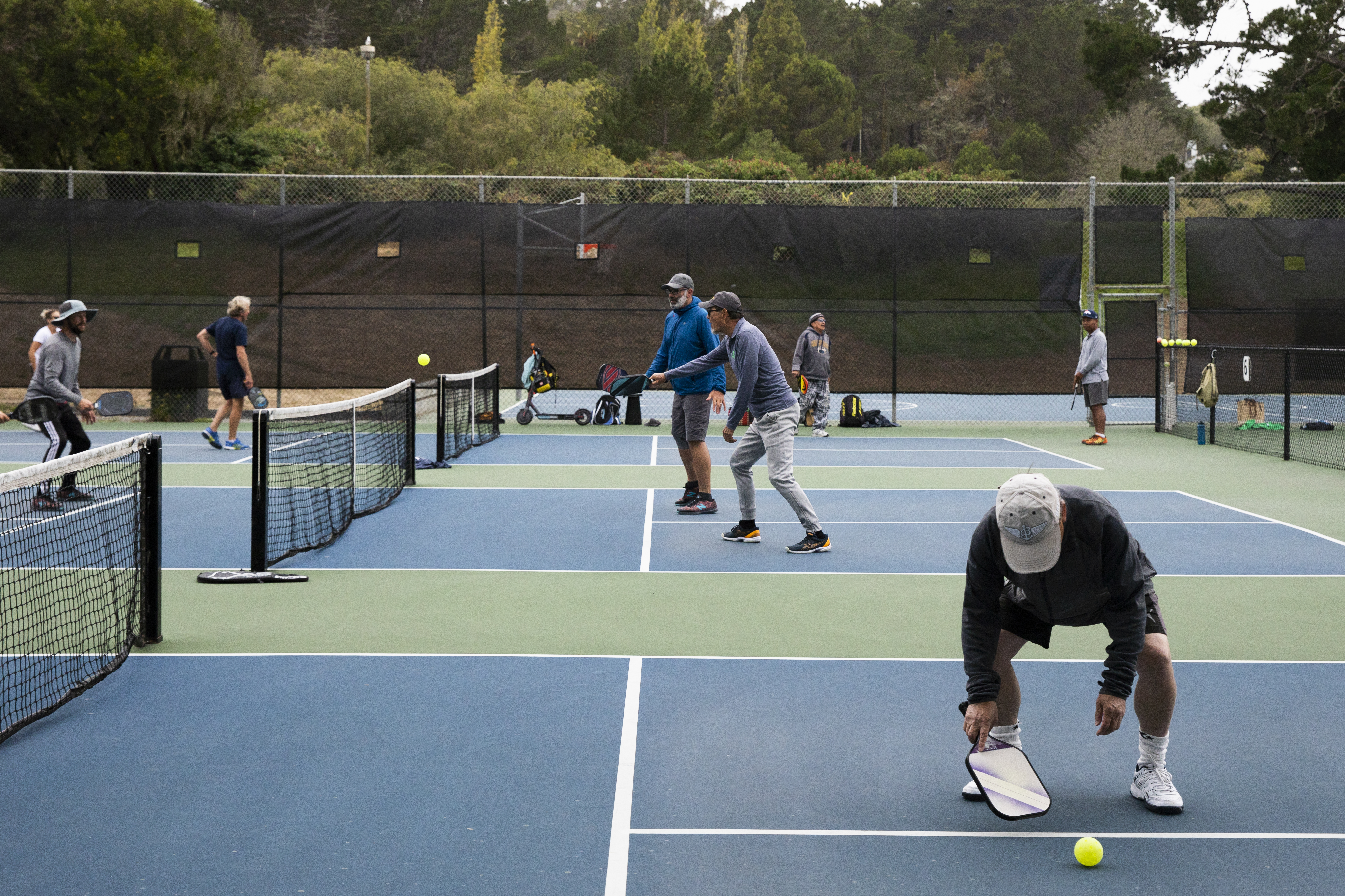 A man bends down on a pickleball court to grab a ball with other players in the background