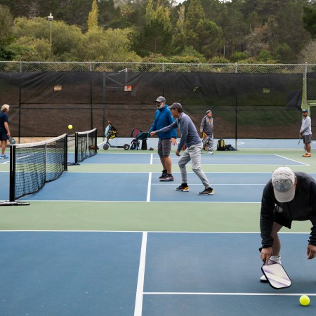 A man bends down on a pickleball court to grab a ball with other players in the background