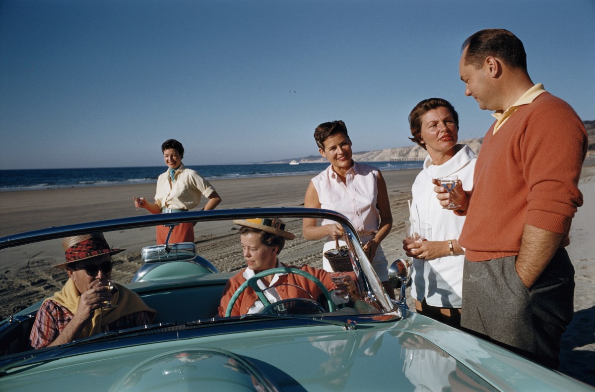 A group of wealthy people gathered around a convertible on a beach.
