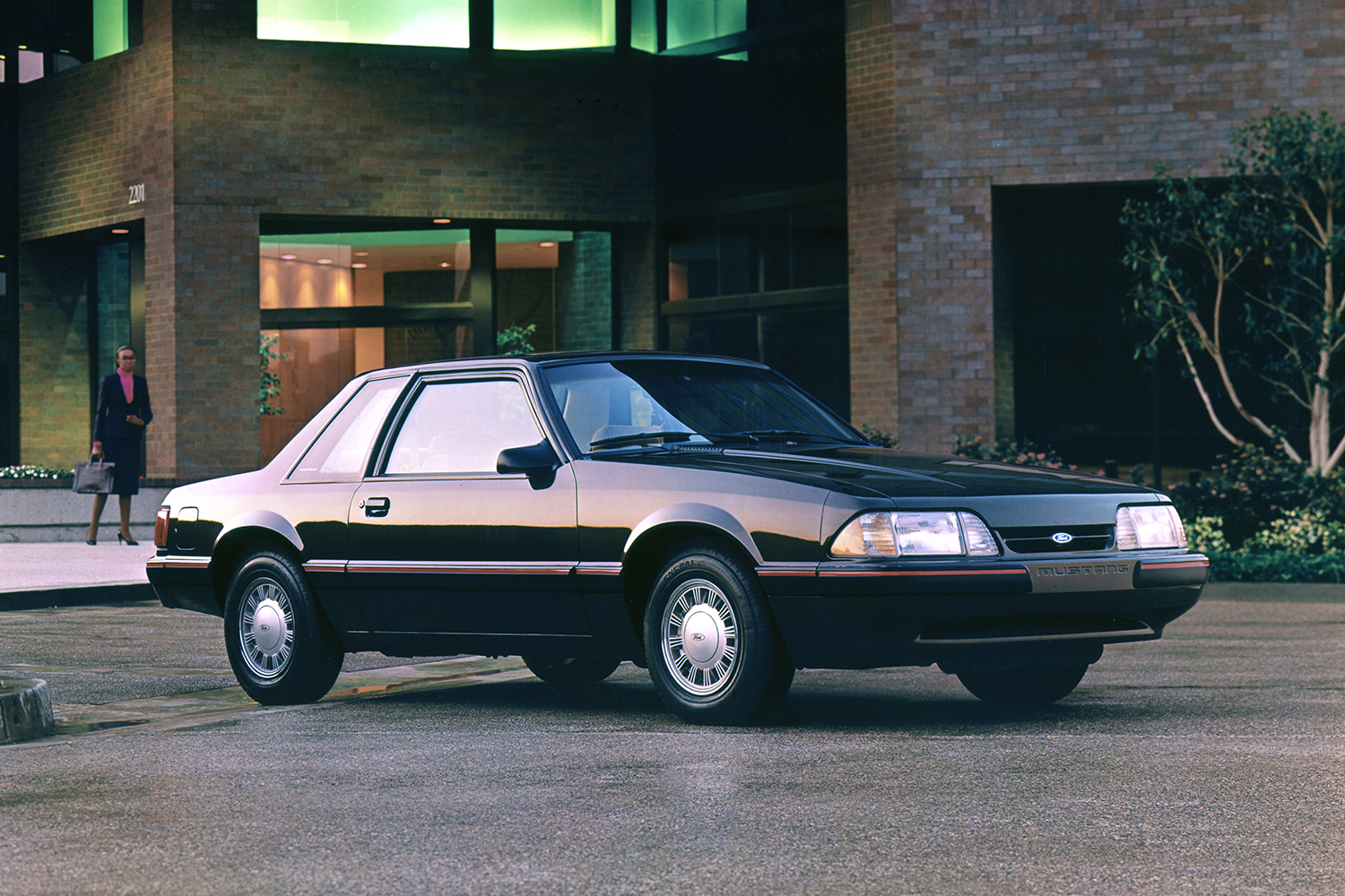 A 1988 Ford Mustang LX with a woman standing in the background.  The Mustang 5.0 is now considered a modern classic car.