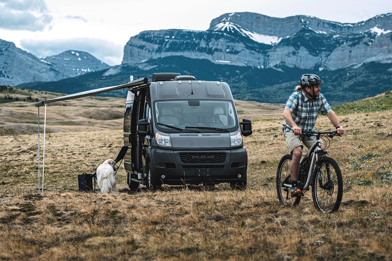 A man cycling away from an Airstream Van in a grassy valley next to the mountains