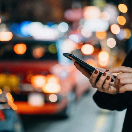 Close up of young woman using mobile app device on smartphone in downtown city street, with illuminated busy city traffic scene during rush hour with traffic congestion in the evening