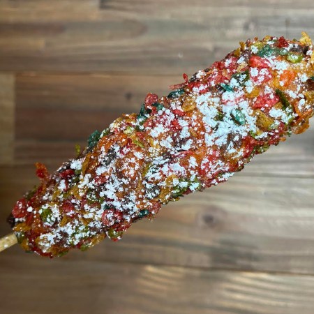 A person holding a Rainbow Kong Dog, a corn dog with fruity rice puffs and powdered sugar. It's part of the sweet hot dog trend.