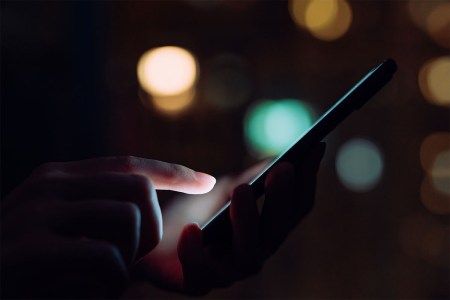 Close up of woman's hand using smartphone in the dark, against illuminated city lights. A tech tool called Fog Reveal uses phone data and can be used to circumvent warrants.