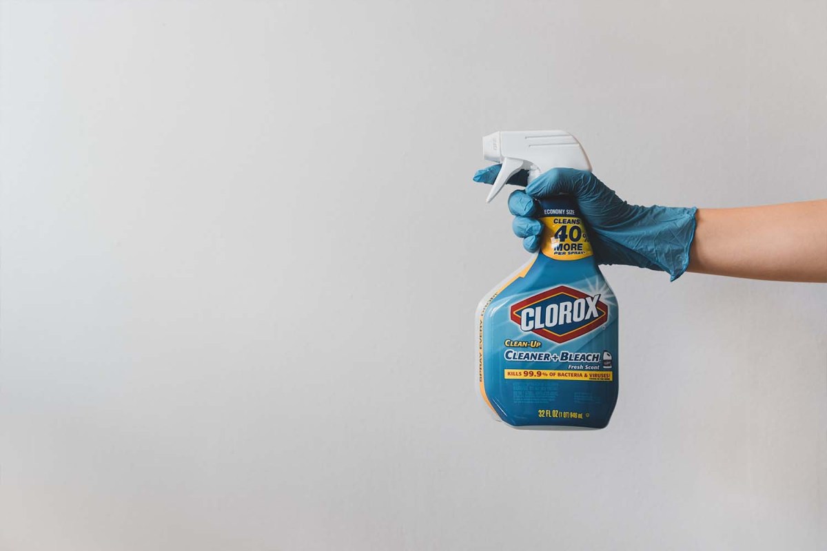 A gloved hand holding a bottle of bleach cleaner against a white background