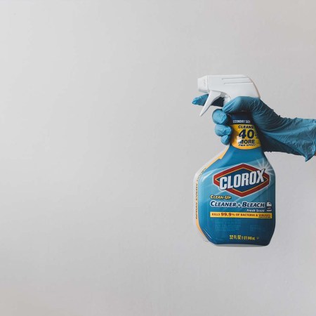 A gloved hand holding a bottle of bleach cleaner against a white background