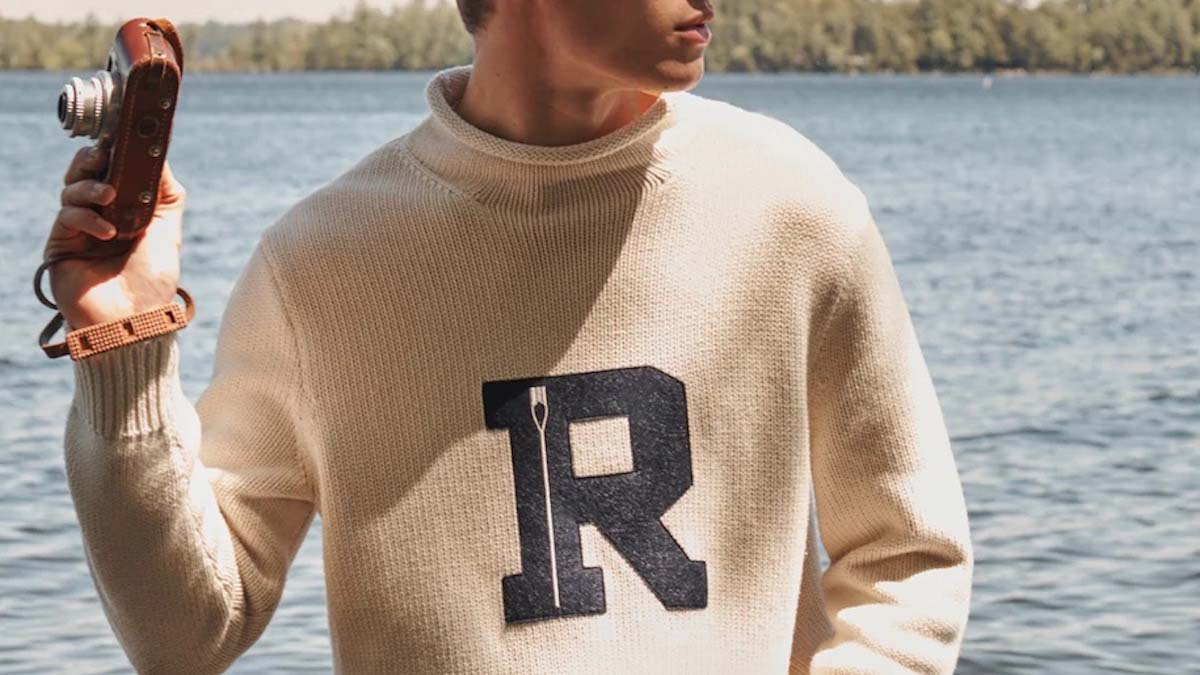 a model holding a camerain a cream cotton rollneck varsity sweater from J.Crew against a lake background