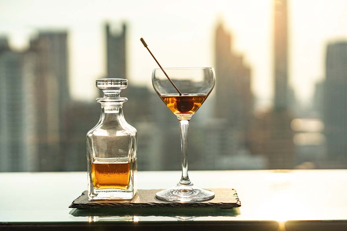 Photo of alcohol in a decanter and a cocktail in a coupe glass overlooking a skyline