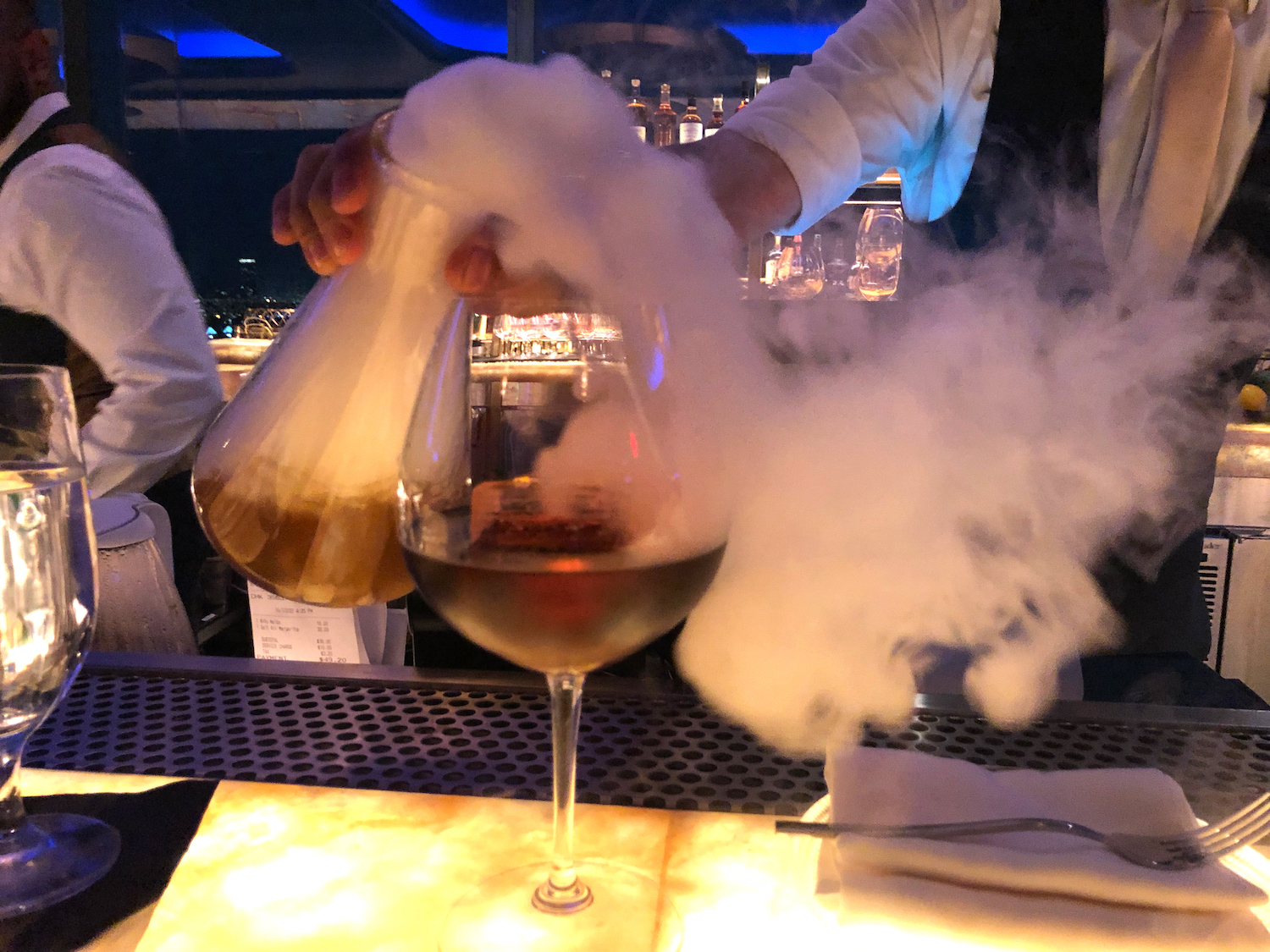 A bartender at Nubeluz pours the Foggy Hill cocktail from a beaker vessel into a wine glass
