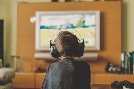 A boy with a headset playing video games on the TV, his back to the camera.
