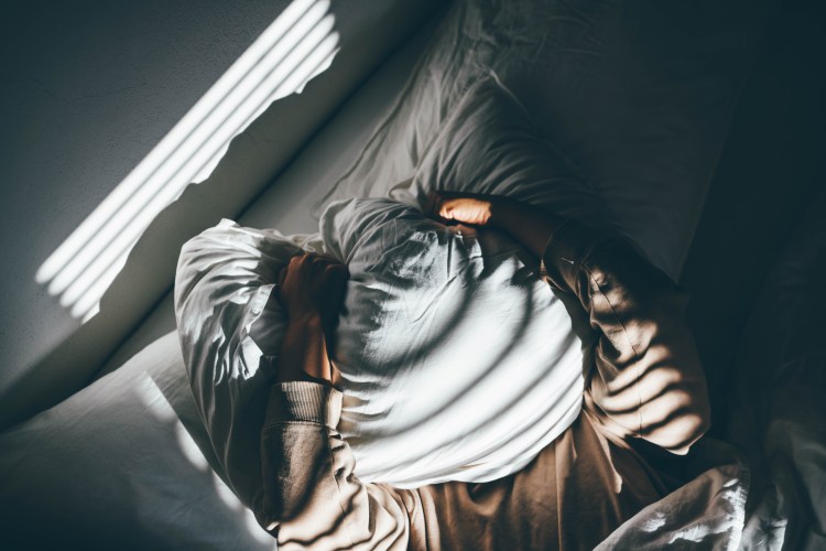 A man holds a pillow over his face as light trickles into his bedroom.