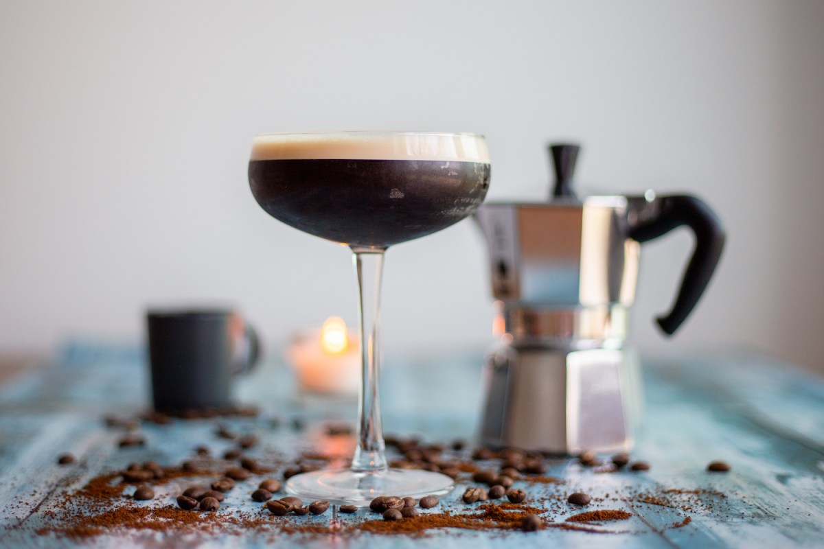 Espresso Martini in a coupe cocktail glass, on a blue wooden table, with blurry images of a mocha pot, candle and an espresso cup