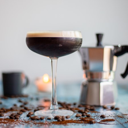 Espresso Martini in a coupe cocktail glass, on a blue wooden table, with blurry images of a mocha pot, candle and an espresso cup