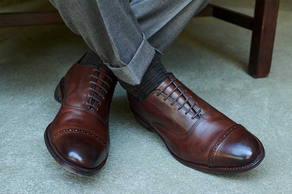 A person crossing their legs wearing a pair of Allen Edmonds dress shoes. The footwear brand is taking 40% off a number of styles before Christmas.