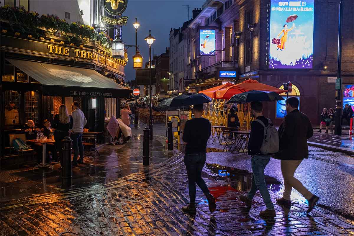 On a rainy night in Soho, Londoners walk across a pedestrianised Old Compton Street. Due to increased public urination, a splashback paint is being applied to various areas around the city.