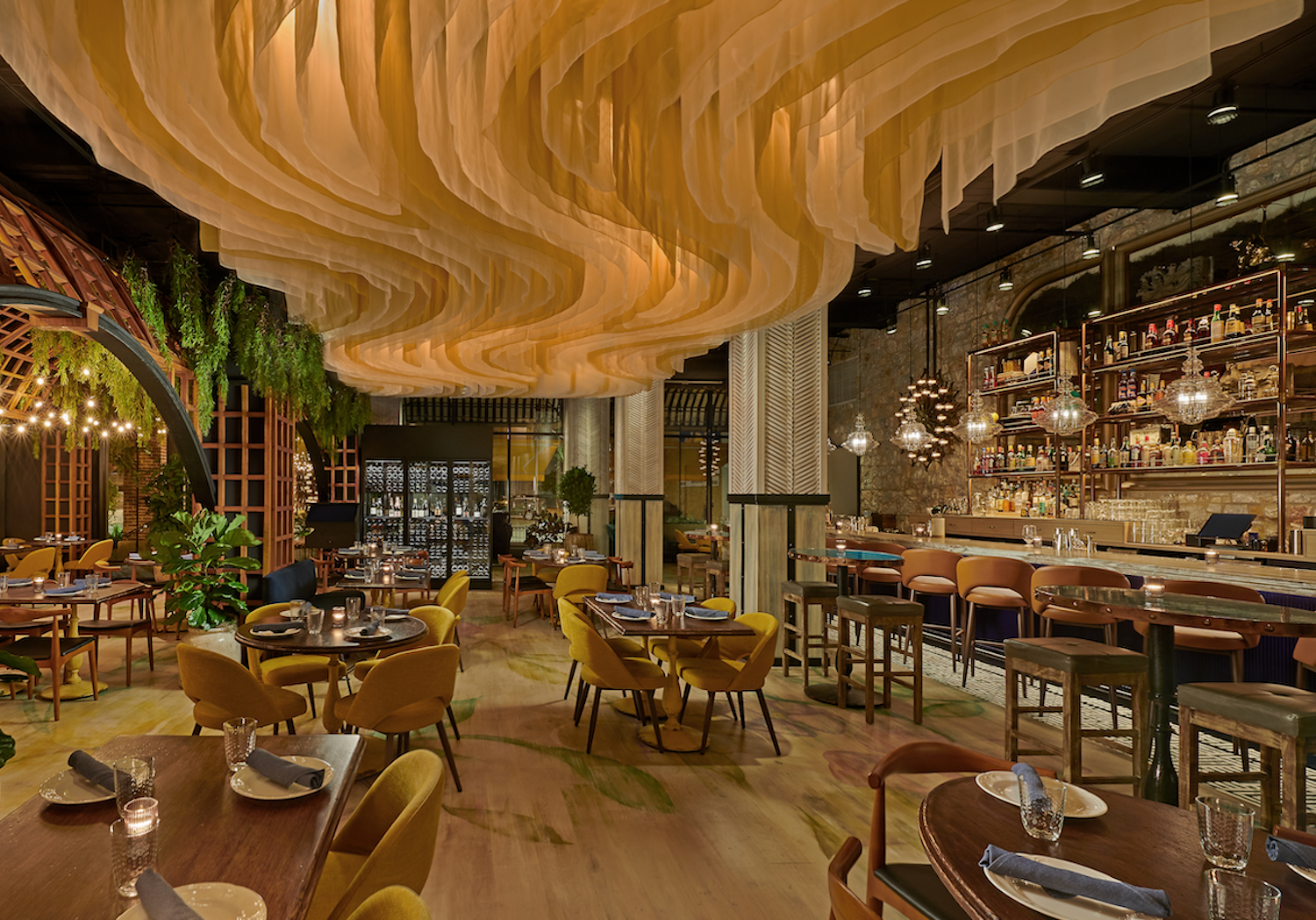 Interior of Alla Vita with yellow chairs, booth seats circular tables and a large bar with high-top seating in a dark brown/wooden/brick wall interior, surrounded by greenery