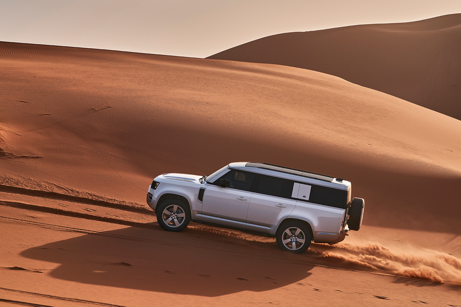 A Land Rover Defender 130 in white driving up sand dunes