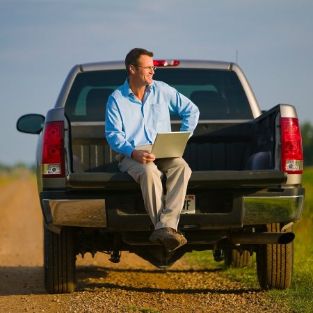 A man in a button-down shirt and khakis sitting in the bed of a pickup truck with a laptop computer. According to survey data, pickup truck buyers know they don't actually need their trucks.