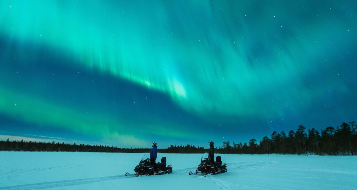 two people standing with their snowmobiles other the northern lights