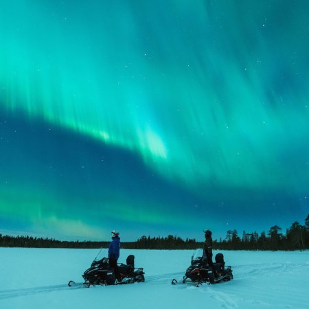 two people standing with their snowmobiles other the northern lights