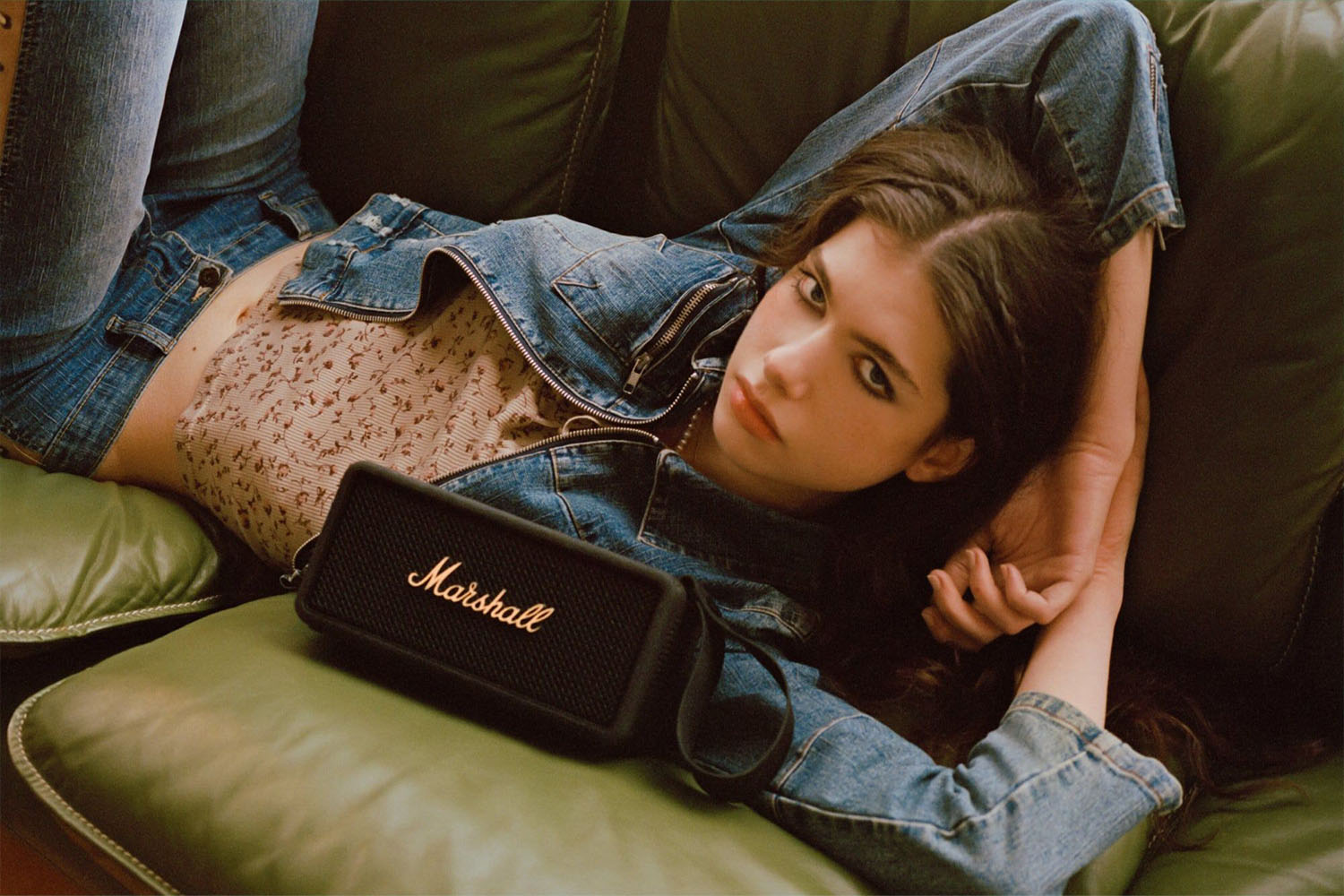 a model laying on a couch next to a black Marshall Middleton Bluetooth Speaker