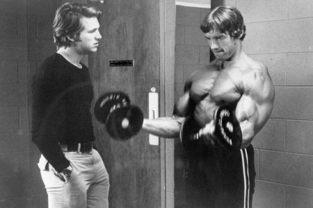 Jeff Bridges talking to a shirtless Arnold Schwarzenegger, who is curling a barbell.
