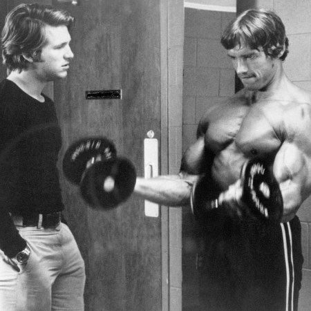 Jeff Bridges talking to a shirtless Arnold Schwarzenegger, who is curling a barbell.