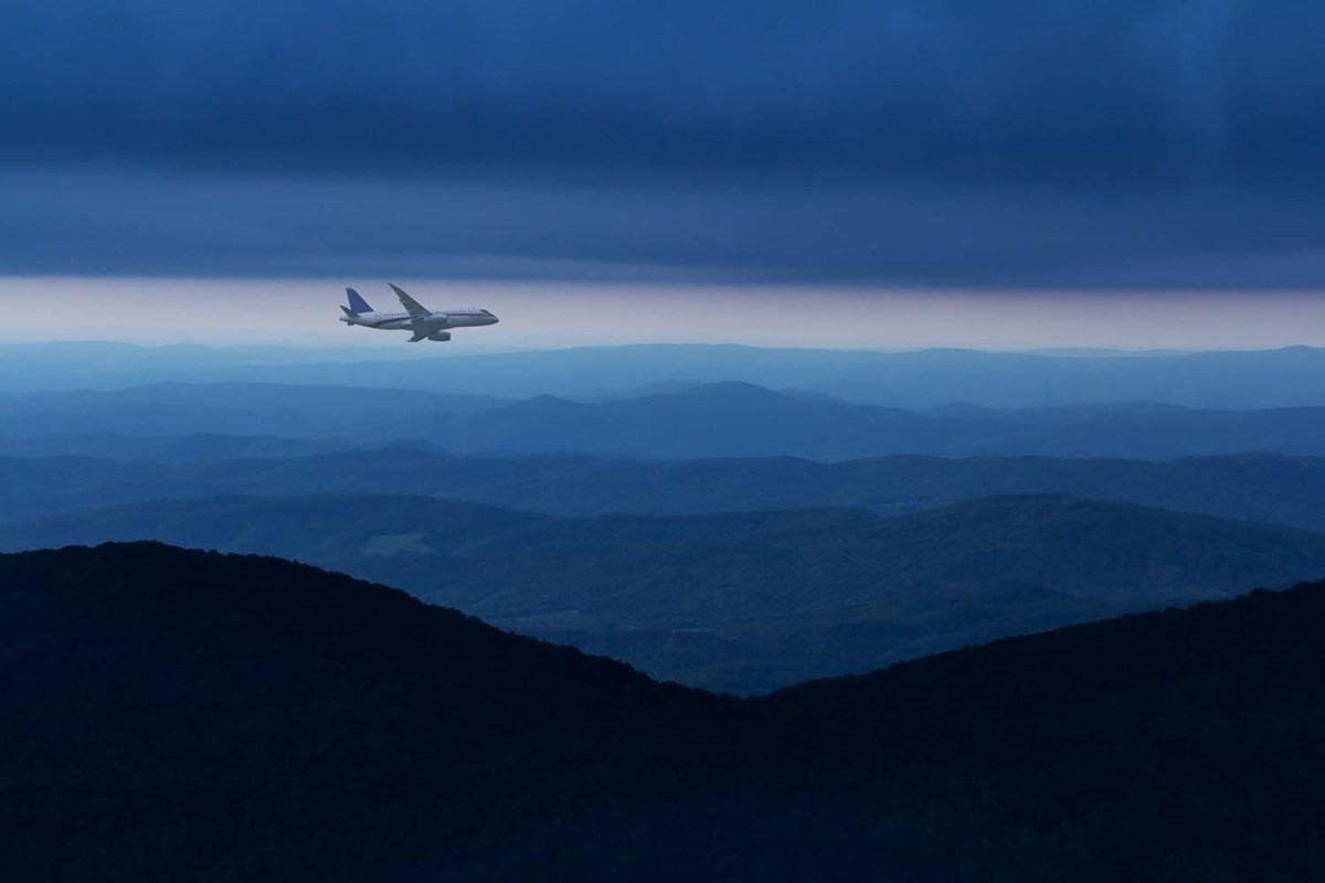 Commercial airplane flying above mountain ranges in cloudy sky at night.