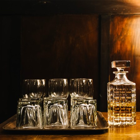 An ornate glass bottle of half empty whisky with a set of glasses and tumblers on a serving tray, in atmospherically lit on an old-fashioned, classy wood-panelled bar.