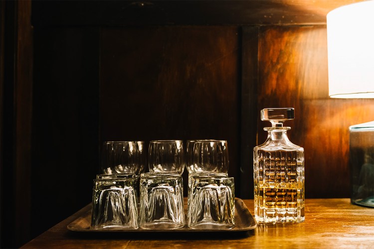 An ornate glass bottle of half empty whisky with a set of glasses and tumblers on a serving tray, in atmospherically lit on an old-fashioned, classy wood-panelled bar.