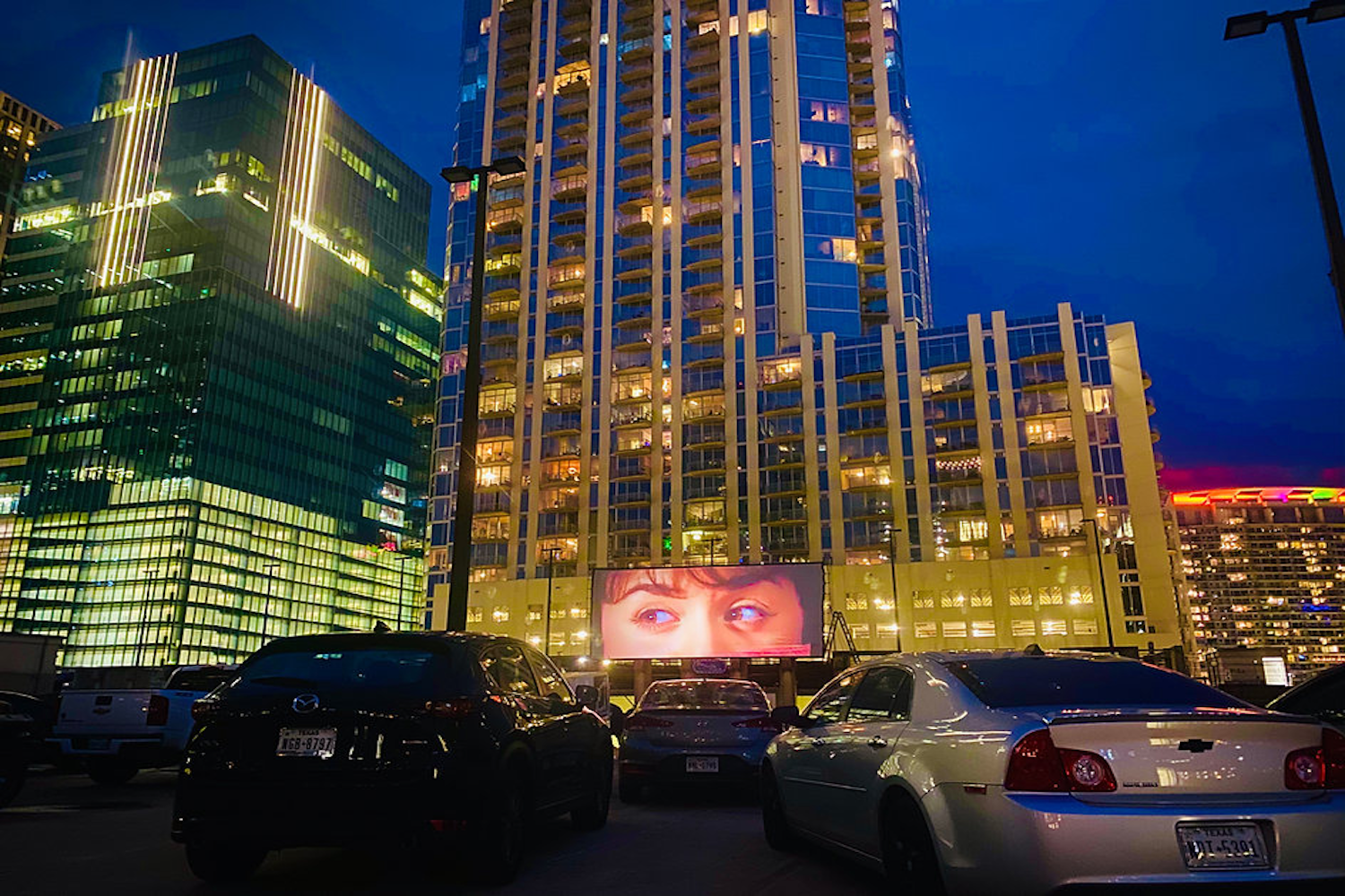 Cars parked by city buildings pointed at a screen playing a movie.