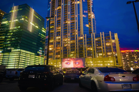 Cars parked by city buildings pointed at a screen playing a movie.