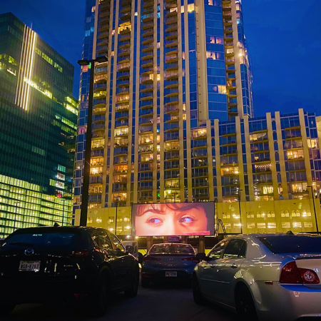 Cars parked by city buildings pointed at a screen playing a movie.