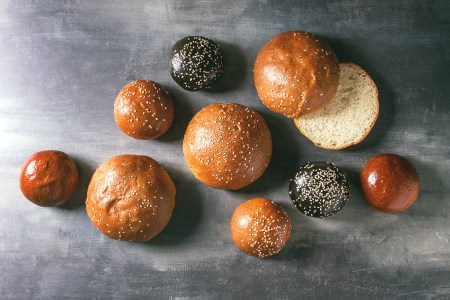 Variety of homemade traditional and black cuttlefish ink different size burger buns with sesame seeds whole and sliced over blue texture background