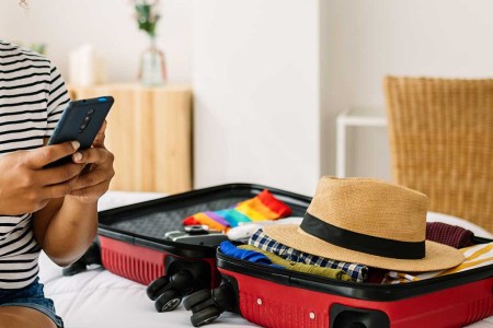 young female tourist hanging cellphone sitting on bed while preparing luggage before going on summer vacation. Expedia just launched ChatGPT in its travel app.