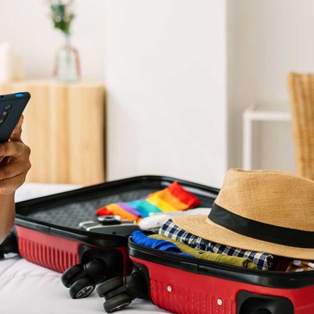 young female tourist hanging cellphone sitting on bed while preparing luggage before going on summer vacation. Expedia just launched ChatGPT in its travel app.