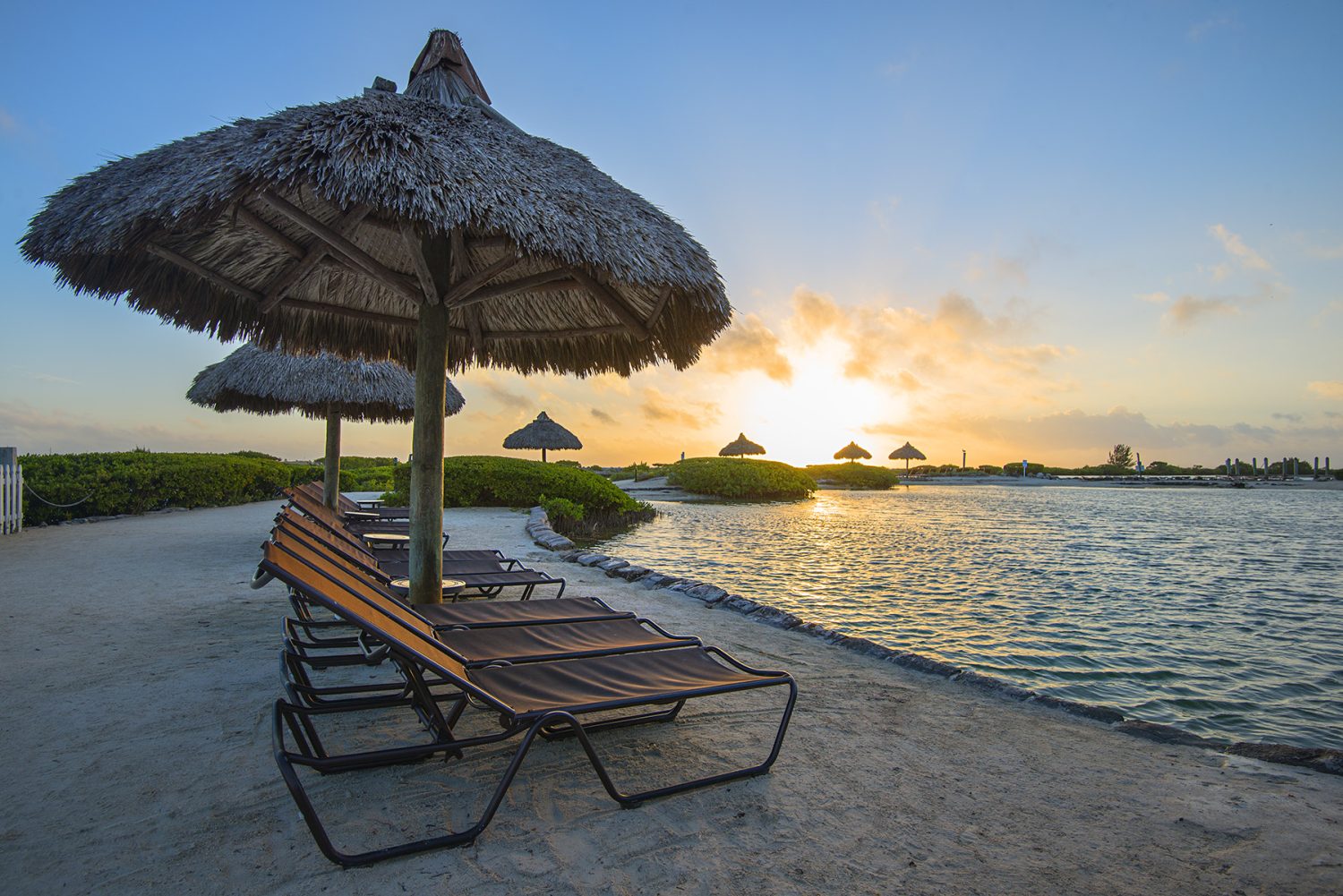 Umbrella and lounge chairs lined up along a lagoon waterfront at sunset.