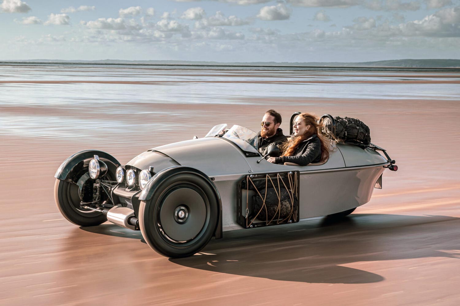 Two people driving in a Morgan Motor Company Super 3 three-wheeled car on the sand