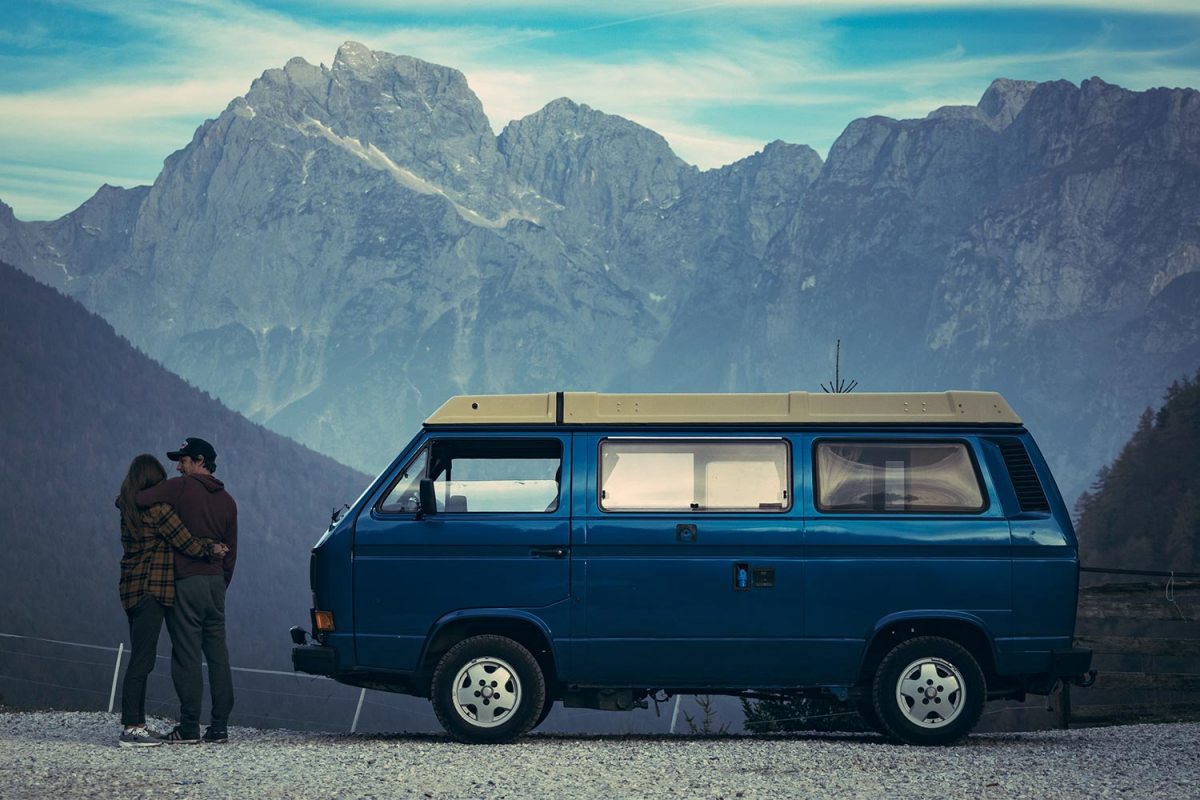 A man and a woman stand next to a camper van with mountains in the background
