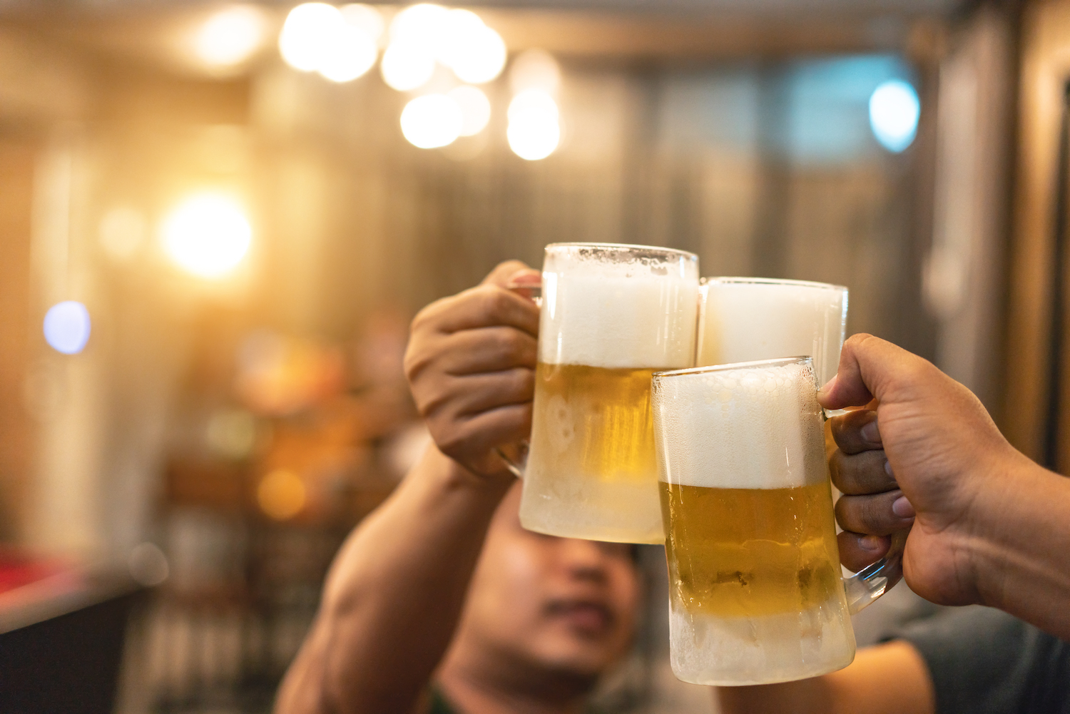 Beer glasses raised in a toast. Close-up hands with glasses. Blurred bar interior at the background
