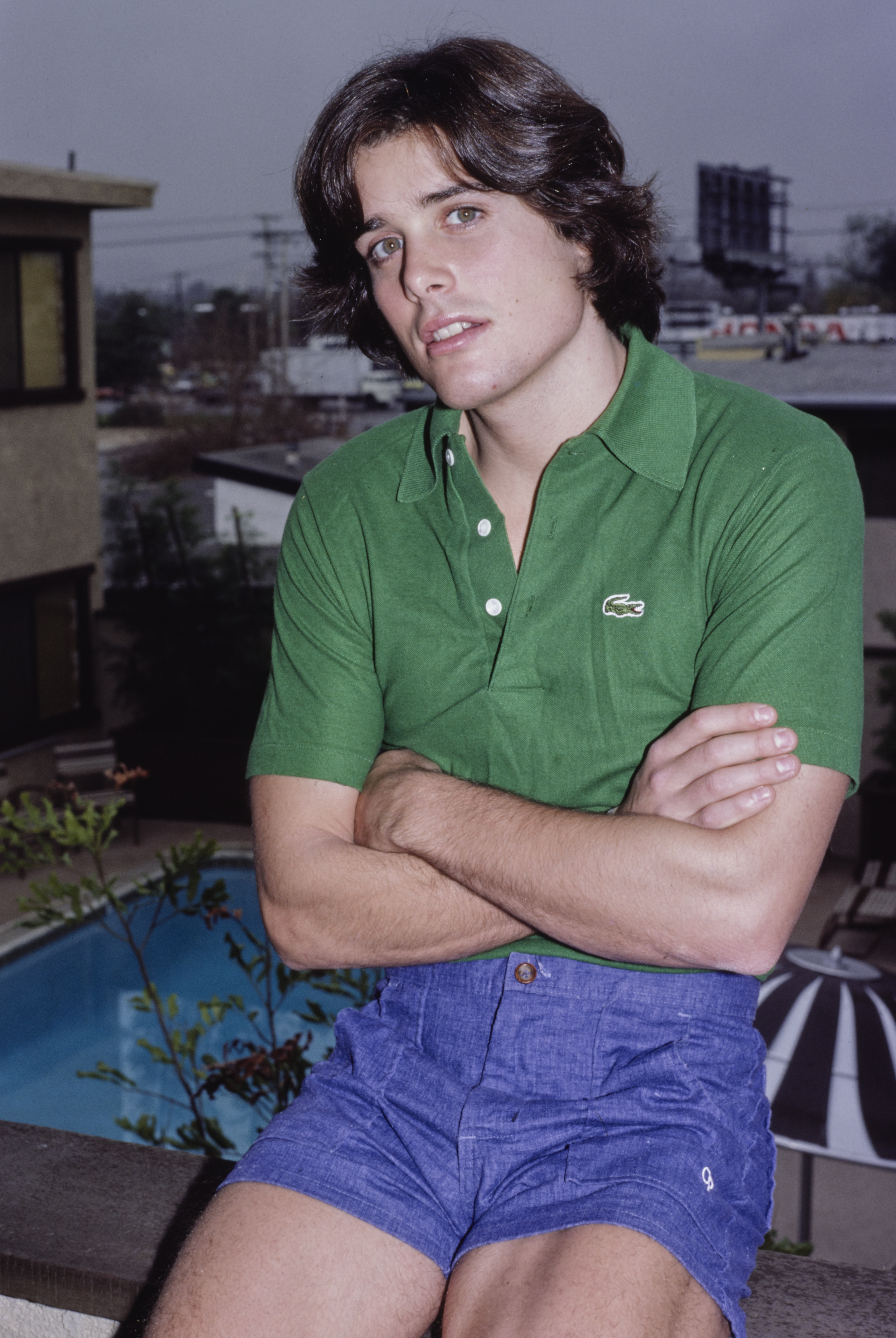 American actor Peter Barton sits, his arms crossed, wearing a green Lacoste polo shirt and blue shorts, a swimming pool below the ledge on which he sits, location unspecified, circa 1980. (Photo by Vinnie Zuffante/Michael Ochs Archives/Getty Images)