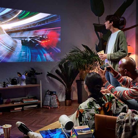 The Freestyle by Samsung in a living room where three people are playing video games. The Discover Samsung Event features discounts across the tech brand's portfolio.