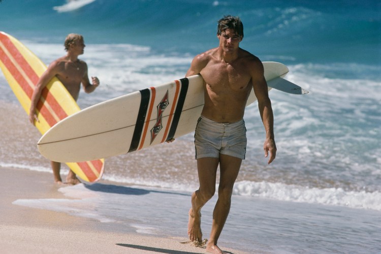 An actor playing a surfer, walking down the beach.