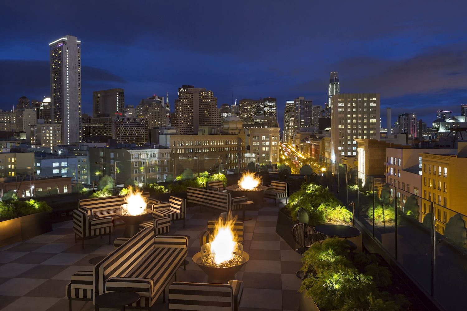 city skyline from a rooftop at night with tables and couch seating and fire pits.