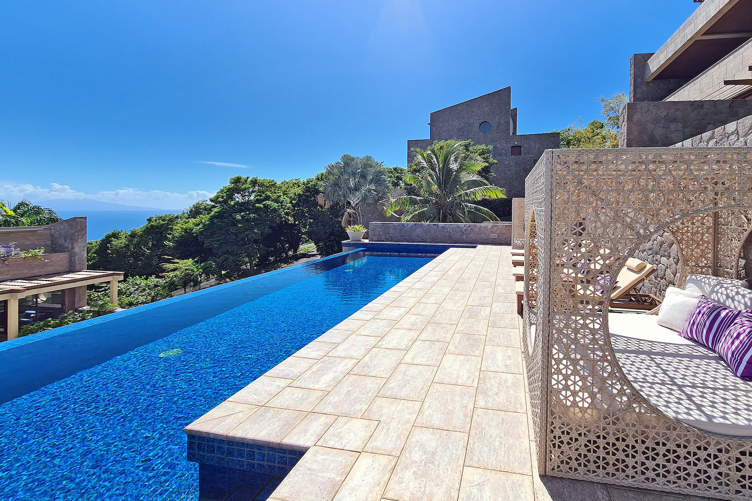 a tiled deck alongside a bright blue pool surrounded by greenery on a sunny day.