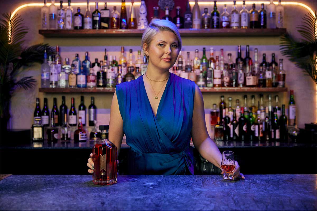 Forbidden distiller Marianne Eaves behind a bar holding a glass and a bottle of Forbidden bourbon
