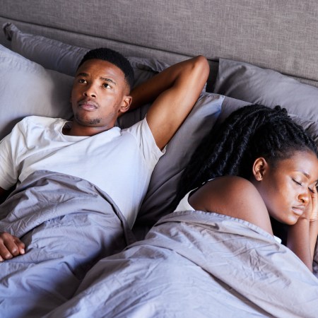 Young man looking unhappy while lying in bed with his sleeping wife