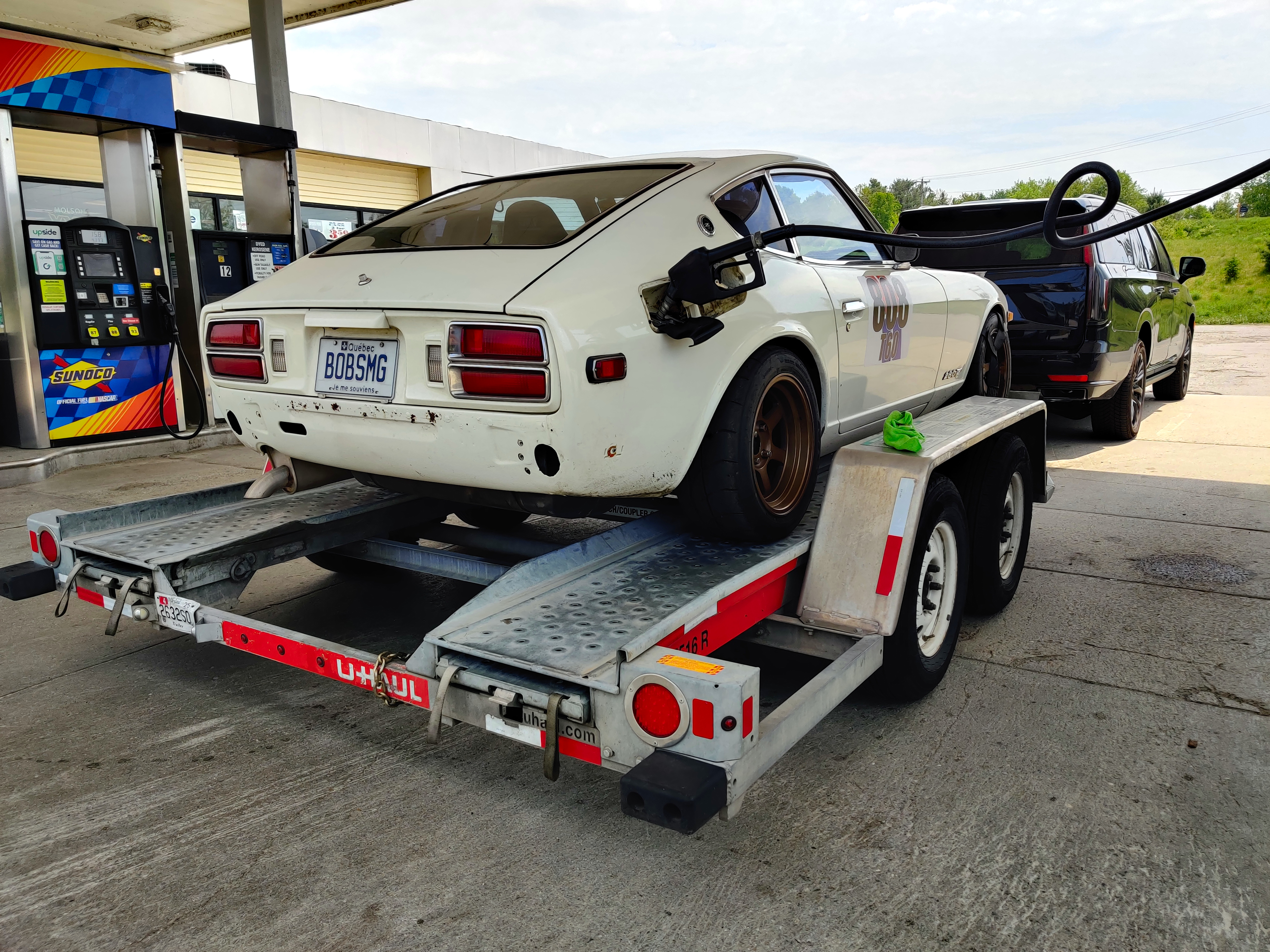 A 1978 Datsun 280Z sits on a trailer in a gas station receiving gas from a pump hose